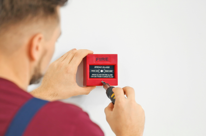 man installing a fire alarm onto a plain, white wall to show why you need to know about the UK fire alarm regulations